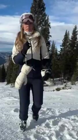 A woman dressed in ski gear, including a white and navy jacket, navy ski pants, a helmet, and goggles, is kneeling on a snowy mountain slope. She is holding a pair of white ski mittens. The camera is positioned from a first-person perspective, presumably held by someone sitting on the snow, wearing red ski pants and ski boots with skis attached. The woman interacts with the camera holder, seemingly engaging in a conversation or demonstration. The background features a scenic view of snow-covered mountains and evergreen trees under a partly cloudy blue sky. The scene appears to be at a ski resort on a sunny day.