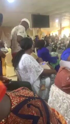 The video shows an indoor gathering of people, likely a party or celebration. People are seated on chairs while others stand and walk around. A woman is holding a baby. Some individuals are dressed in formal or traditional attire. The room appears to be a hall or event space with decorated elements. The people seem to be enjoying themselves.