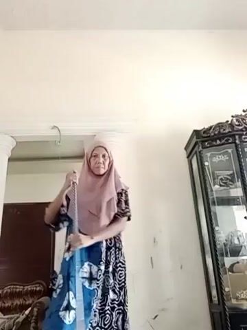 A woman wearing a pink hijab and a patterned dress is standing in a room, holding a mop. She is looking towards the camera and appears to be in the process of mopping the floor. The room has light-colored walls and a decorative display cabinet on the right side of the frame. There are also pillars and a glimpse of furniture in the background. The lighting suggests the video was filmed indoors during the daytime.