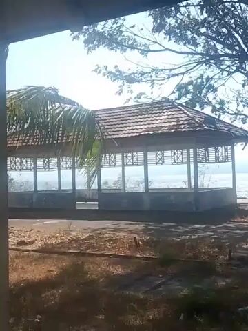 The video captures an outdoor scene dominated by a shaded, open-air pavilion. The pavilion has a tiled roof and is supported by concrete pillars. Intricate latticework is visible on the walls of the pavilion. Beyond the pavilion, a body of water, likely the ocean, can be seen under a bright, clear sky. The foreground of the video is filled with dry, sparse vegetation and some debris, suggesting a somewhat unkempt environment. The overall impression is one of a scenic overlook or rest area that may be disused or in need of maintenance.