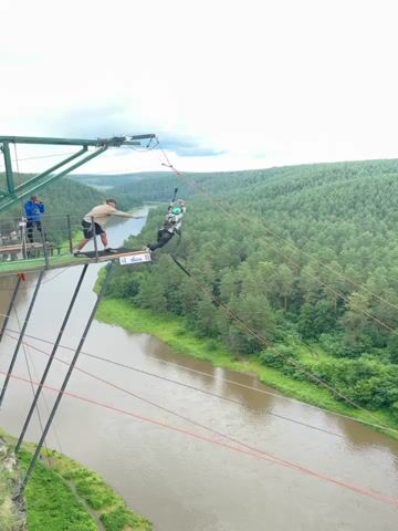 The video captures a bungee jump from a platform overlooking a wide, muddy river flanked by dense green forest. In the foreground, a person is mid-air, attached to a bungee cord, their body in a dynamic pose as they descend. They are wearing a patterned jacket, dark pants, and a helmet. Two individuals are visible on the platform at the start of the jump. One, wearing a light brown jacket and shorts, stands near the edge, looking down. Another person in a blue jacket is further back.

The scene is set in a natural, outdoor environment, likely a park or recreational area given the presence of the bungee jumping setup. The river flows through a valley, with steep, forested banks rising on either side. The trees are lush and green, suggesting a summer or late spring setting. The sky appears overcast, with diffused light, indicating it might be a cloudy day.

The primary activity is the bungee jump, a thrilling adventure sport. The perspective shifts from a wide shot showing the jump and the surrounding landscape to a closer view of the river and its banks as the jumper descends. The overall mood is one of excitement and adrenaline, inherent to the activity.

There is some visible text on a white sign attached to the platform, but it is too small and blurry to be legible. The location context provided is Kul'metovo, Russia, which aligns with the natural, forested landscape.