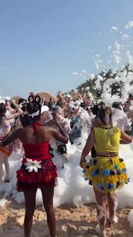 The video captures a lively beach party scene with a large group of people dancing and having fun amidst a foamy, bubbly atmosphere. The foam appears to be generated by a machine, creating a surreal, snow-like effect on the sand and beachgoers. Two individuals in the foreground, one wearing a red outfit and the other a yellow outfit, are facing away from the camera, with their backs to the viewer. They are both adorned with decorative skirts, the one in red featuring white floral accents, and the one in yellow decorated with blue heart-shaped embellishments. The crowd is energetic, with many individuals raising their hands in the air, suggesting a celebratory mood. Palm trees are visible in the background, along with a clear blue sky, indicating a sunny day. The overall impression is one of vibrant celebration and enjoyment at an outdoor event.