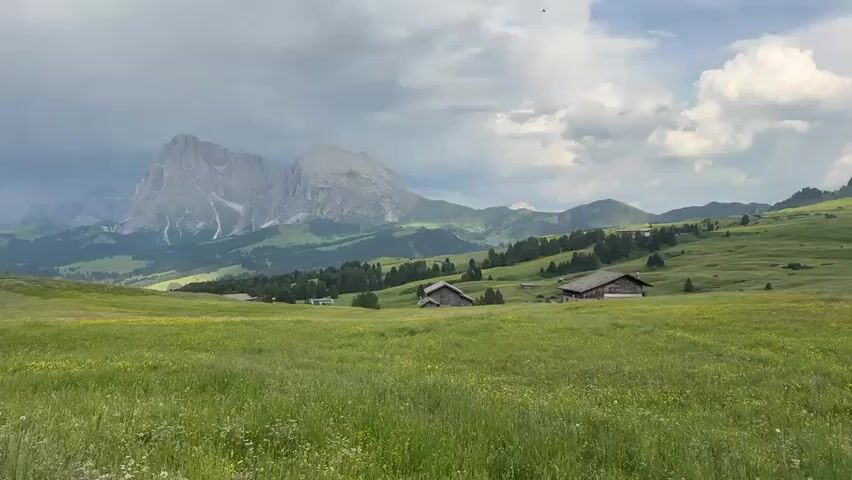 This is a video showcasing a scenic landscape of the Dolomites in Kastelruth - Castelrotto, Italy. The scene is dominated by rolling green hills covered in lush grass and wildflowers, with several small wooden chalets nestled into the landscape. In the background, imposing mountain peaks rise majestically, their rugged forms contrasting with the softer slopes. The sky is partly cloudy, with dramatic formations of white clouds against a backdrop of grey, suggesting it might be a day with changing weather, possibly late afternoon or early evening.

The overall impression is one of tranquility and natural beauty. The absence of people suggests a peaceful, uninhabited environment, emphasizing the grandeur of nature. The wooden chalets, with their traditional architecture, add a touch of rustic charm and hint at human presence in harmony with the natural setting. The vibrant green of the meadows and the scattered yellow wildflowers create a cheerful and inviting atmosphere. The scale of the mountains evokes a sense of awe and wonder. The video appears to be a continuous panning shot across this breathtaking vista, allowing the viewer to take in the panoramic views. There is no visible text in the media file.