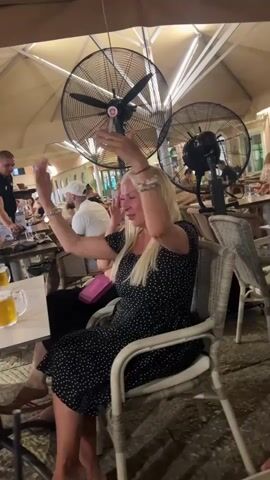 Two women are sitting at a table outdoors. The woman on the left is wearing a black dress with white polka dots and has blonde hair. She is raising her hands in the air and smiling. The woman on the right is wearing a black dress with white polka dots and has dark hair. She is looking at the woman on the left and smiling. There are two large fans mounted above them, and other people are visible in the background sitting at tables. A table with drinks is in the foreground.