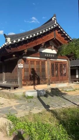 The video pans across a traditional Korean courtyard on a sunny day with a bright blue sky and scattered clouds. The foreground features green grass and a paved stone pathway. On the left, a traditional Korean wooden building with a tiled roof is visible. It has intricate wooden lattice doors and windows. A sign with Korean characters is mounted above the entrance. A shadow of a person taking the video is cast on the building. To the right of the building, a wooden utility pole with a sticker showing a number and Korean characters stands in front of some flowering bushes. The camera then pans further to the right, revealing more of the courtyard. More traditional Korean buildings with tiled roofs are visible in the background, along with trees. A signpost with Korean text is also present. The video then moves to a wider view of an open, paved area with several benches and large umbrellas providing shade. In the center of this area, a person in a dark dress is standing and holding a camera, appearing to take photos. Further in the background, other people and structures are visible, including what looks like a modern building with a dark tower. The scene appears to be a historical or cultural site, possibly a village or a museum. The overall atmosphere is peaceful and sunny.