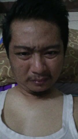 The video shows a man lying on a bed, looking directly at the camera. He is wearing a white tank top. A brown and white patterned pillow is behind his head. The man's expression is mostly neutral, with slight eye movements. The background suggests a bedroom setting. The lighting is somewhat dim.