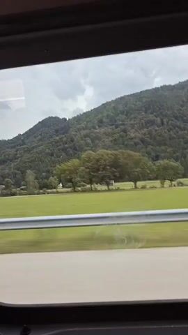 The video shows a view from a train window looking out at a rural landscape. The landscape consists of rolling green hills covered in dense trees, a flat green field, and scattered buildings, including what appears to be a church spire with a red roof on the far left. There are also some deciduous trees in the middle ground. A silver guardrail runs horizontally across the lower portion of the scene, separating the field from a road or track below. The sky is overcast with dark clouds. The camera is moving, indicated by the slight blur and the shifting perspective of the landscape. The view is framed by the dark interior of the train and the window.