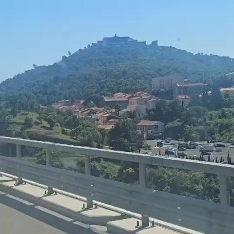 The video shows a view of a town and a large fort on top of a hill, taken from a moving vehicle, likely a car, that is traveling on a bridge or elevated road. The foreground features a silver-colored guardrail. Below, there's a partially visible parking lot, suggesting a potential rest stop or viewpoint. A small town is nestled at the base of the hill, comprising houses with terracotta roofs. The hill itself is densely covered with green trees. At the top of the hill, a large stone fort stands prominently against the light blue sky. The camera zooms in to show the architecture of the fort, including its fortifications and a tower structure. The sky is clear and bright, indicating a sunny day.