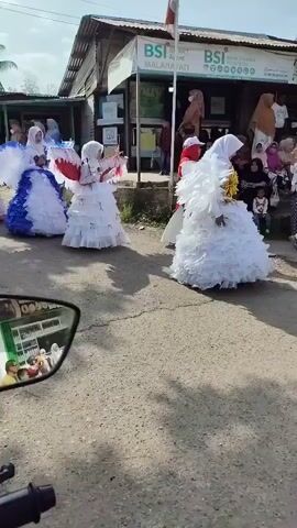A group of young people, possibly children or teenagers, are participating in a parade or procession. They are dressed in elaborate costumes, resembling angels, with large white wings and flowing gowns in various colors, including white, blue, and pink. Some are also wearing white headscarves. The procession is taking place on a dirt road in front of a building with signage that appears to be a bank, possibly "BSI" or "BSI Malahayati". There are spectators on the sidewalk observing the event. The video is captured from the perspective of someone on a motorcycle, with the motorcycle's side mirror reflecting a portion of the scene, including some of the spectators. The weather appears to be sunny with a clear sky. The overall atmosphere seems festive and celebratory.