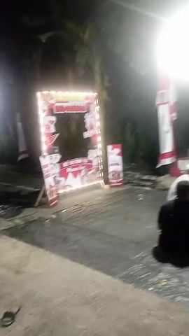 The video captures a nighttime outdoor scene. In the foreground, several people are seated on the ground, which is covered with a dark tarp. One person in the center appears to be interacting with small objects on the tarp. To the left, a decorated photo booth or backdrop is visible. In the background, a small elevated structure with a red roof, possibly a kiosk or stall, is set up. Several people are seated or standing around this structure. A bright, large light source illuminates the area, creating strong shadows. Flags with red and white patterns are displayed on poles next to the kiosk. The overall atmosphere suggests a gathering or event taking place after dark.