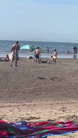 A group of people are on a sandy beach. A man stands on the left, facing away from the camera. Three young boys are playing with a soccer ball in the center. One boy is kicking the ball, another is running, and the third is kneeling. In the background, several other people are in the ocean, some swimming and some wading. Beach umbrellas and towels are visible. The sky is clear and blue, and the water is calm.
