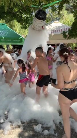 A group of children and adults are playing in a large pile of white foam outdoors. The foam appears to be generated from a machine at the top of the frame, which is a large white cylindrical object suspended from above. People are dressed in swimwear and appear to be enjoying themselves, interacting with the foam and each other. The environment is an outdoor setting with trees and some structures in the background that suggest a recreational area or park. The sun is shining, indicating daytime.
