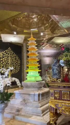 The video shows a brightly lit, ornate interior, possibly a temple or a luxurious room. The central focus is a tall, multi-tiered pagoda sculpture that glows with green and yellow light at its base and transitions to orange and yellow towards its apex. The pagoda stands on a tiered, marble-like pedestal. To the left of the pagoda, a large black panel with an intricate gold tree-like design is visible, along with white orchids in a pot. To the right, there is a decorated altar or display table with a blue and white vase, flowers, and other ornate objects, including what appears to be a small shrine. The ceiling is adorned with elaborate gold patterns and lighting. The overall impression is one of opulence and a spiritual or decorative setting.