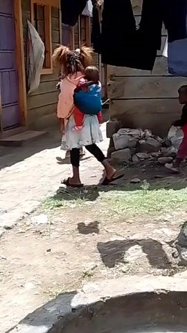 The video shows a group of young children walking along a narrow path outside a building. One child is carrying a baby on her back in a blue carrier. Another child, wearing red, is walking alongside the first child. A third child is walking behind them. The building is light brown with purple doors and window frames. Laundry is hanging on a clothesline in the background. The path is cracked and uneven, with patches of grass and a small concrete structure visible. The scene is shot from a slightly elevated position.