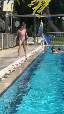 A young girl in a colorful swimsuit is on the edge of a swimming pool, appearing to prepare for a jump. She is positioned on the paved deck beside the bright blue water, her body bent in a crouched stance. Her hair is pulled back, and she wears goggles. Nearby, a man is in the pool, facing away from the camera, with his head and shoulders above the water. In the background, there are trees, a fence, lounge chairs, and a blue plastic chair. Yellow and blue pennants are strung overhead, suggesting a festive or recreational atmosphere. The scene appears to be outdoors on a sunny day, judging by the bright lighting and shadows. The girl's posture and the man's presence indicate a swimming or pool activity. There is text visible on a white sign in the background, but it is too small and pixelated to read. The location is identified as Beit Yizhaq - Shaar Hefer, Palestinian Territories.