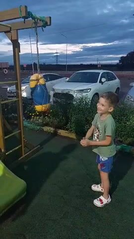 A young boy is punching a punching bag that is hanging from a wooden frame outdoors. The boy is wearing a green t-shirt and blue shorts. The punching bag is yellow and blue and shaped like a Minion character. The boy is standing on a green rubberized surface. Behind the boy, there are several cars parked. The sky is overcast with some clouds, suggesting it is either dusk or dawn. There are also some plants and bushes in the background.