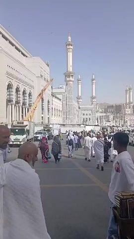 A wide shot reveals a busy city street in Mecca, Saudi Arabia, thronging with people. Many of the individuals are clad in white garments, signifying their participation in a pilgrimage or religious activity. Prominently featured in the background is the Masjid al-Haram, or Great Mosque of Mecca, with its distinctive white minarets reaching towards the sky. Adjacent to the mosque, the Abraj Al-Bait Towers rise majestically, including the iconic clock tower. Construction cranes indicate ongoing development in the area. Various vehicles, including taxis and private cars, traverse the road, highlighting the urban setting. The camera follows the movement of people walking across the street, focusing on the visual elements of the scene.