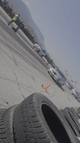 The video shows a drag race taking place on a paved track. Two cars, a white BMW wagon and a silver sedan are lined up at the starting line, where a race official dressed in a yellow vest stands ready to signal the start. A crowd of spectators lines the side of the track, beyond a series of orange cones. The track is marked with tire marks from previous races. The tires and a hand holding a mobile phone are visible in the foreground. In the background, there is a mountain range with a partially cloudy sky. After the start, the cars accelerate down the track.