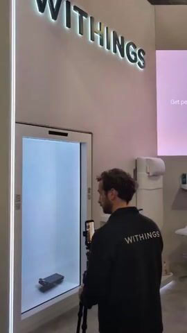 The video shows the Withings booth at an event.  The Withings sign is visible.  There is a large display screen showing a woman standing on a Withings scale. The display screen reads, "Get personalized longevity guidance." Several people are at the booth. One person is wearing a Withings jacket.  Another sign reads "Withings Body Scan 2." There is a demonstration table with products.