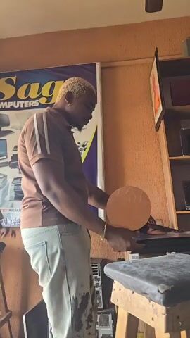 A light-skinned man is standing inside what appears to be a computer repair shop. He has blonde, curly hair, a beard, and is wearing a brown polo shirt with white stripes on the sleeves, light blue jeans with dark stains, and a gold bracelet. He is holding a round, golden-colored object with a patterned surface in his left hand and using his right hand to operate a piece of equipment on a workbench. The workbench is covered with a worn, black pad. Behind him, there's a sign that says "Sage COMPUTERS" and a shelf with various electronic devices. The wall is painted a tan color and has a framed picture hanging on it. There is a wooden stool next to the bench. Loud music plays in the background.