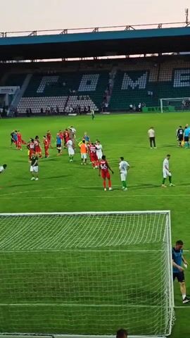 The video shows a football stadium with players on the field. Spectators are visible in the stands, which have green seats with white lettering that appears to spell out "HOME". Several players in red and green uniforms are gathered on the field, seemingly at the end of a match. A person in a blue shirt and black shorts is near a goal net, appearing to be a ground staff member. The sun is setting, casting a warm light over the scene.