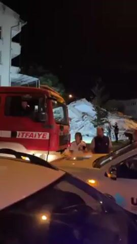At night, a red and silver fire engine with the word “İTFAİYE” on it, which means “Fire Department” in Turkish, is parked on the side of a street lined with buildings. There are a number of people, many of whom are men, standing around on the street. Behind the fire engine, a large pile of rubble is visible, suggesting a possible building collapse or some other kind of destruction. The video seems to be taken from inside a car looking out onto the scene. The sound of the fire engine and possibly other emergency vehicles can be heard in the background.