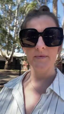 A woman wearing large, dark sunglasses and a white pinstripe shirt stands outdoors. Her hair is pulled back in a bun, and she is wearing hoop earrings and a necklace. She looks directly at the camera with a slight smile. In the background, there are trees and some industrial-looking structures, possibly water tanks. The lighting suggests it is daytime.