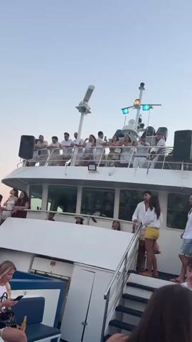 The video shows a boat party at dusk. Several people are visible on the boat, some standing and others seated. The boat is white with multiple decks and appears to be moving through the water. In the background, there is a coastline with hills and buildings. The sky is a pale blue and orange, indicating sunset. The video is shaky and blurry in some parts, suggesting it was filmed on a moving platform.