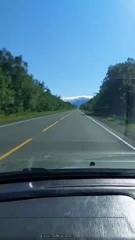 The video shows a first-person perspective from inside a vehicle driving on a paved road. The road is flanked by lush green trees and vegetation on both sides. The sky is clear and blue, with a few wispy clouds near the horizon. In the distance, a mountain range is visible, its peaks partially obscured by clouds. The road appears to be a two-lane highway with a yellow dividing line and a white shoulder line. The dashboard of the vehicle is visible in the foreground, and there is a small black object on the dashboard. The video captures the scenery as the car drives forward.