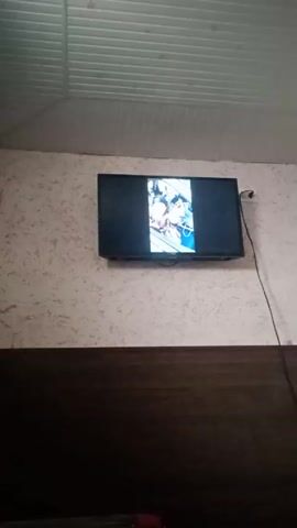 A wall-mounted television displays what appears to be a comic book scene. The TV is mounted on a wall that is light colored with a mottled pattern. Below the textured wall is a dark brown wood panel. Above the TV is a white drop ceiling. A power cord for the TV hangs down. Music can be heard.