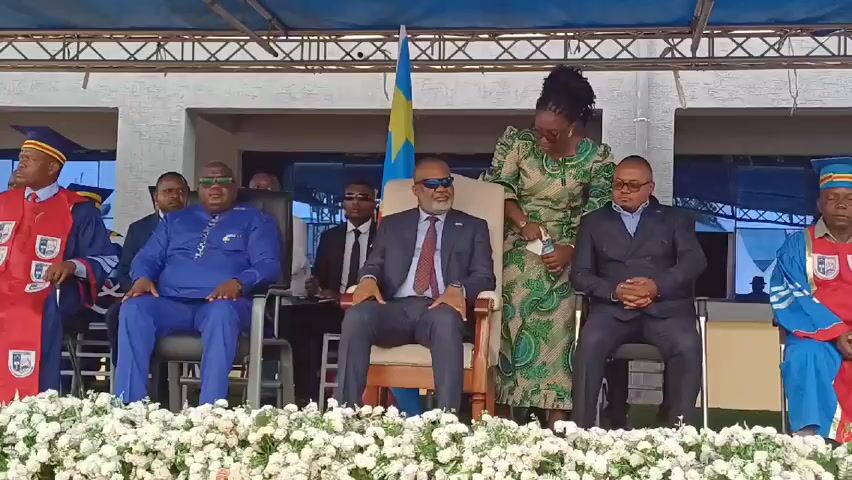 On a stage, nine people are visible, five seated in the front row. From left to right, the first person is wearing a red and blue academic gown and cap. The second is wearing a blue dashiki with green sunglasses. The third is wearing a black suit, red tie, and blue sunglasses, seated in a beige chair. The fourth person is wearing a green patterned dress and appears to be adjusting the suit of the person to their left. The fifth person is wearing a dark suit and glasses. Flanking the group on the right is a person in a blue academic gown and cap. Behind the seated people, there are four additional figures in suits and ties, wearing sunglasses. The flag of the Democratic Republic of the Congo stands behind the seated figures. A structure with a blue tarp overhead and metal latticework runs across the top of the frame. A row of white flowers decorates the front of the stage.