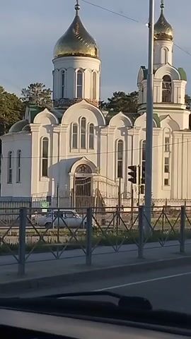 The video shows a white church with gold domes from the perspective of a moving car. The church is behind a gray fence. A traffic light is visible. The sky is light blue. Trees are visible in the background. The car is moving. Music is playing.