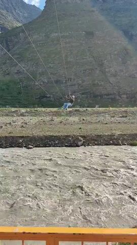 The video shows a person zip-lining across a muddy river. The zip line is suspended between two tall mountains. The person appears to be a young male wearing a helmet and a harness. The river is wide and turbulent, with strong currents. The surrounding landscape is mountainous and barren, with sparse vegetation. There are some structures visible on the riverbank, including what appears to be a small building and some recreational equipment. The weather appears to be clear and sunny.