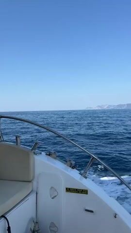 A video taken from the perspective of someone on a moving boat. The ocean is visible with light waves and a clear blue sky. In the distance, land with mountains can be seen. The front of the boat is visible in the foreground, with a seat and metal railings. There is a yellow and black warning sticker on the side of the boat.
