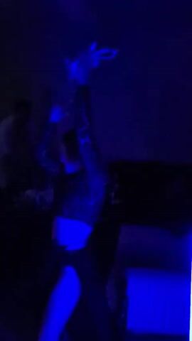 A short video shows a woman dancing in two different settings. Initially, she's indoors under blue lighting, wearing a leopard print top and dark skirt with a high slit. Another person is dimly visible in the background. The second scene shifts to an outdoor setting, possibly a rooftop or patio, again under blue lighting. A partially constructed building is visible. The woman is now in a light-colored, likely white, dress or cover-up and dancing with two men. A laptop is also visible in the foreground, possibly playing music. Overall, the video seems to depict a party or social gathering with dancing and music.