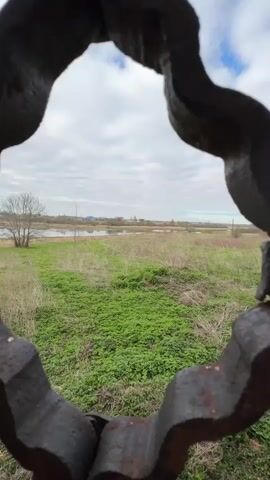The video opens with a view through an ornate, dark metal grate, revealing a rural landscape beyond. Green fields stretch towards a body of water, possibly a river or lake, with distant buildings under a cloudy sky. The camera then pulls back slightly, showing more of the intricate, scrolled metalwork set into an old brick wall. A close-up shot focuses on the crumbling brick wall, where plaster is peeling away, exposing weathered red bricks and deteriorating mortar. The final shot is an interior view of a ruined brick building, characterized by multiple arched openings and remnants of a ceiling structure. Much of the roof is missing, allowing the cloudy sky and a tree branch growing through the opening to be visible. The brickwork throughout the structure is old and in disrepair, with patches of plaster clinging to the walls. The scene depicts the historical ruins in Staroe Rakomo, Russia, on an overcast day.