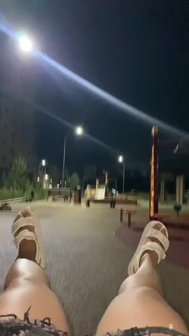 The video shows a first-person perspective of someone sitting on a playground at night. The person is wearing ripped denim shorts and white sandals. The playground is illuminated by streetlights, casting long shadows. In the background, there are playground structures, benches, and buildings. The camera is pointed downwards, focusing on the person's legs and feet. The person appears to be stationary or moving very slowly. The overall atmosphere is calm and quiet, with a sense of solitude.