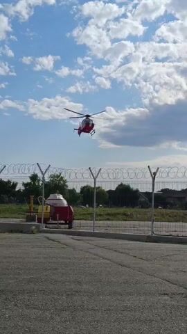 A red and white helicopter flies in a blue sky with scattered clouds. The helicopter is in the upper half of the frame, angled slightly downward. In the foreground, there is a chain-link fence with barbed wire at the top. Behind the fence, to the left, is a red and yellow wheeled cart with a white, dome-shaped object on it. The ground in front of the fence is paved, and further in the background, there are trees and some buildings. The sun appears to be shining, casting light on the scene.