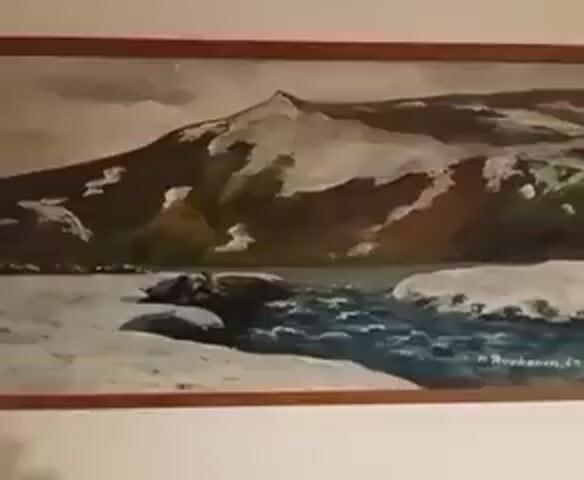 A painting of a mountain landscape with snow and a river is displayed on a wall. The painting is framed in wood. The camera slowly pans across the painting.