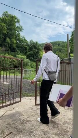 Three young men, dressed in white polo shirts and black pants, walk through an open metal gate. The man in the foreground is carrying a large, thin binder with a purple and white cover. The gate appears to lead to a dirt or gravel driveway. Beyond the gate, lush green trees and a sloping hill covered in vegetation are visible. A yellow building can be seen on the hillside in the distance. The sky is cloudy, suggesting it may be an overcast day. The scene appears to be a rural or semi-rural area, possibly a residential property. The men are walking away from the camera, and their expressions are not visible, but their posture suggests they are moving with purpose. The overall impression is one of casual movement through an outdoor setting.