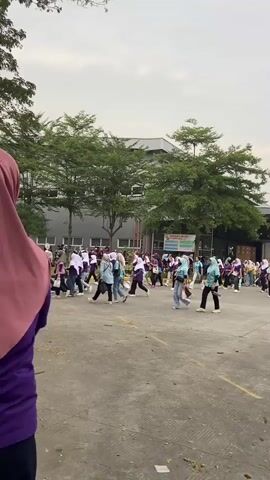 A group of people, predominantly women wearing hijabs and athletic attire in shades of purple and teal, are gathered outdoors in a paved area. They appear to be participating in a group activity or exercise. Some are walking, while others are in motion, possibly stretching or dancing. The background features a modern building with large windows and a row of trees. The sky is overcast, suggesting a temperate climate. The scene is well-lit, and the focus is on the individuals and their collective movement.