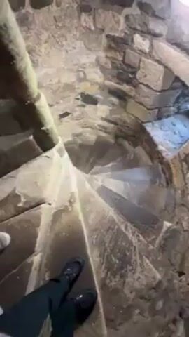 A person is walking down a narrow stone spiral staircase inside a stone structure. The person is wearing black pants and black shoes. The staircase is made of stone and the walls are also made of stone. The lighting is dim. The camera is pointed downward. The person is walking at a steady pace.