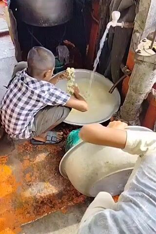 Here's a factual description of the video:

Two children are cleaning what appears to be cooked noodles in a metal bowl.  A boy wearing a plaid shirt and dark pants is crouched next to a large silver bowl that's being filled with water from a tap. He uses his hands to stir the noodles in the water. Another child is partly visible in the foreground, holding a similar empty bowl, seemingly ready to receive the cleaned noodles. The area seems cramped and utilitarian. There are large metal cooking pots visible, suggesting a cooking or food preparation setup. The flooring is stained and has signs of wear.