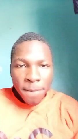 A young Black man is featured in a short video clip, filmed indoors. He has short, dark hair and a faint mustache. He is wearing a bright orange t-shirt.

In the initial frames (0-0:01), he is positioned facing the camera, appearing to sit or stand. He has a relatively neutral expression, with his lips slightly parted, and his right hand is partially visible in the foreground, near his chest.

Around the one-second mark (0:01-0:02), his expression shifts slightly to a subtle smirk, as he continues to look directly at the camera.

From 0:02 to the end (0:04), the man is seen reclined, possibly lying down, with a patterned pillow behind his head. The pillow is brown with white "LV" monograms. He displays a wide, cheerful smile, showing his teeth, suggesting a happy and relaxed mood.

The background throughout the video is a plain, light blue-green wall. There are no other specific objects or activities discernible apart from the man's changing expressions and posture. The visible text is the "LV" monogram on the pillow.
