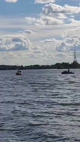 Two individuals are on a paddleboard in the middle of a body of water. Another person is on a jet ski to the right. The sky is cloudy.