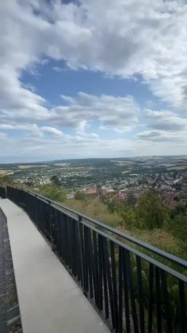 The video shows a scenic view from an elevated vantage point overlooking a town nestled in a valley. A dark metal railing runs along the foreground, separating the viewer from the town below. A paved walkway is visible to the left of the railing. The town appears to be a mix of residential buildings and some older structures, possibly historical, with a church spire or tower visible amongst the rooftops. Lush green trees and vegetation surround the town, and the landscape extends into the distance, with rolling hills and fields under a partly cloudy blue sky. The camera appears to be stationary, capturing a wide panoramic view.
