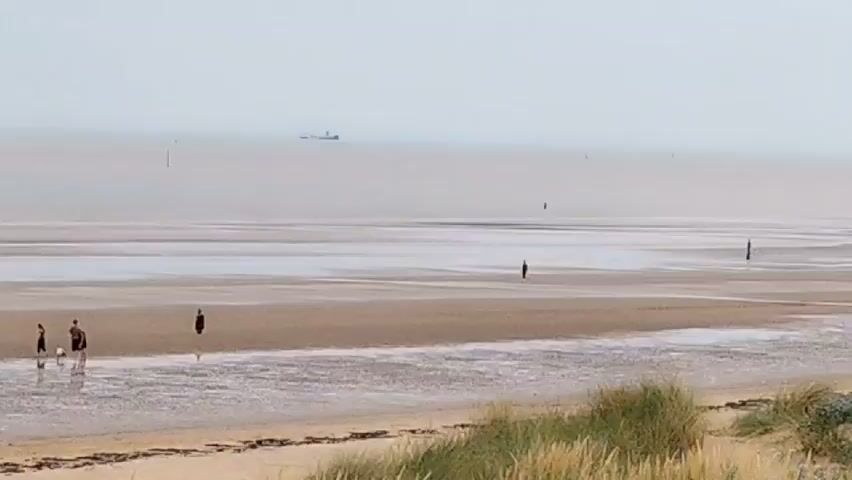 The video shows a wide, flat beach under an overcast sky. The tide is out, revealing a vast expanse of wet sand with shallow pools of water. In the distance, a large ship is visible on the horizon, and several buoys or markers are scattered across the water. On the beach, there are a few scattered figures. In the foreground on the left, a group of three people, possibly a family, are walking. Further back, two lone figures are visible, one standing and another walking. In the far distance, another lone figure stands on the wet sand. The foreground of the beach has dry sand and tall, dry grasses growing in dunes. The overall impression is of a quiet, somewhat desolate coastal landscape.