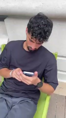A man with curly dark hair, a beard and mustache, wearing a dark t-shirt, is seated on a green chair in front of a light-colored building wall.  He is holding a phone in his hands and looking at it, then turns his head up.  A hand reaches in and pulls his cheek sideways, distorting his face.  He looks at the camera and smiles weakly.