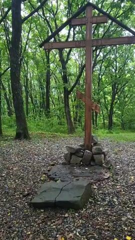 The video shows a wooden cross in a forest clearing. The cross is set on a mound of stones, with a flat rock in front of it. To the left of the cross, there is a wooden bench and a trash can. The trees in the background are tall and covered in green leaves. The ground is covered with gravel and fallen leaves. The video pans slowly to the left, revealing more of the forest and the bench.
