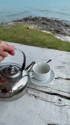 A close-up shot shows a hand holding a stainless steel teapot and pouring hot water into a white teacup sitting on a white saucer. The cup is filled with a light-colored liquid and some loose tea leaves are visible floating in it. A spoon rests on the saucer next to the cup. The scene appears to be outdoors, with a wooden table in the foreground, likely painted white and showing signs of age and weathering. In the background, there is a grassy slope leading down to a rocky coastline and the ocean. The water is a muted turquoise color and appears calm. The sky is a pale blue and overcast.